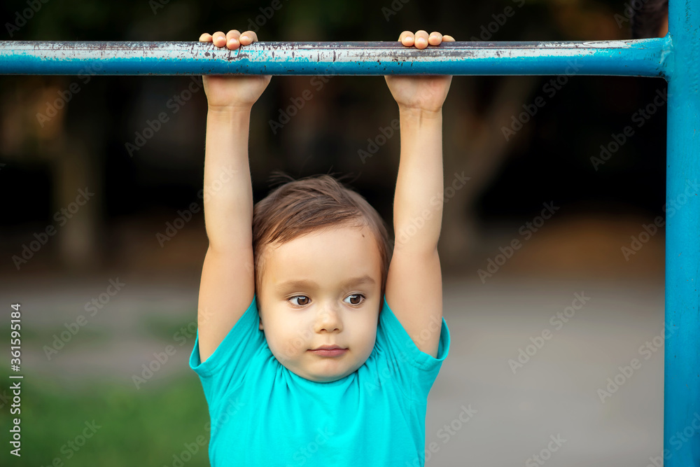 Toddler boy learning to do pull ups at gym lesson in kindergarten: preschooler hanging on worn-out steel bar at sports ground outdoors. Kids and sports concept Toddler boy learning to do pull ups at gym lesson in kindergarten: preschooler hanging on worn-out steel bar at sports ground outdoors. Kids and sports concept