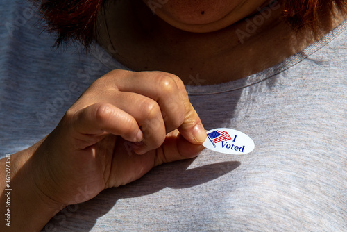 female voter places red white and blue oval I Voted sticker on shirt with right hand after casting ballot
