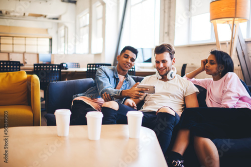 Best friends of millennials sitting on sofa enjoying free time during coffee break and watching funny video in social networks on modern smartphone using free 4G internet connection in coworking space