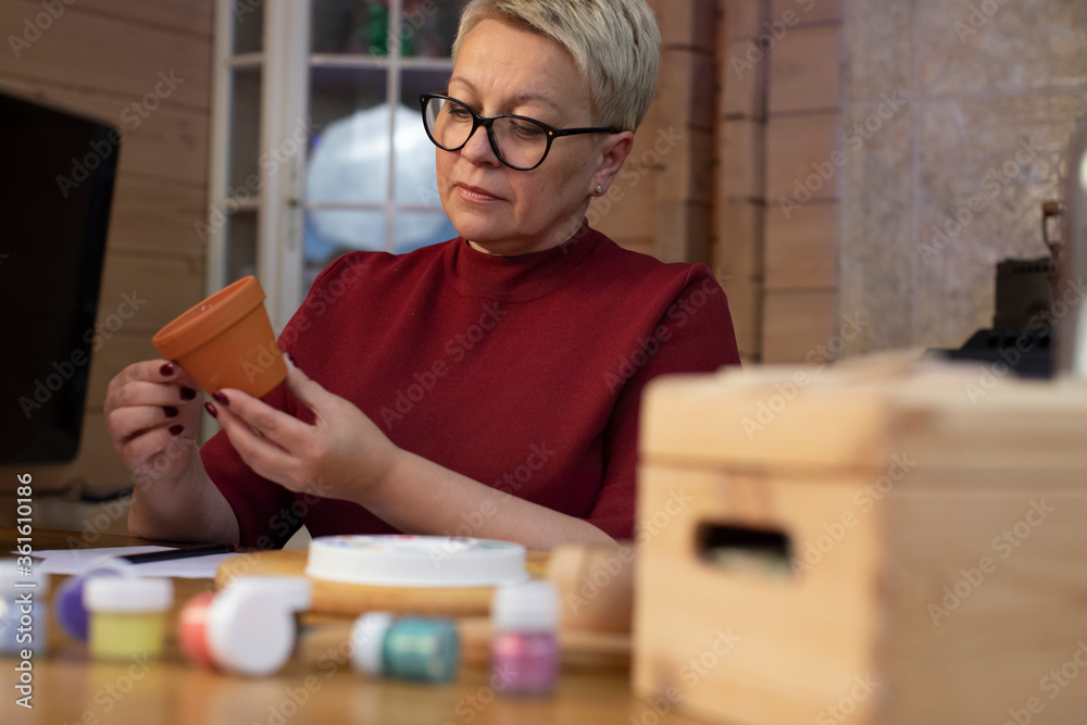 attractive middle-aged woman paints flower pot in cozy country house table view. Concentrated woman with blond hair dressed casually painting. indoors free time concept. Handmade concept attractive middle-aged woman paints flower pot in cozy country house table view. Concentrated woman with blond hair dressed casually painting. indoors free time concept. Handmade concept