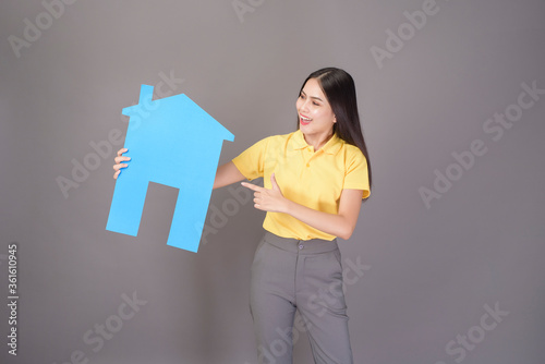 young confident beautiful woman wearing yellow shirt is holding a home singn on grey background studio photo