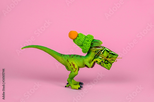 cute green plastic toy dinosaur wearing knitted hat and holding present box on a pink background
