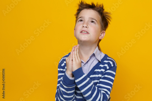 A little funny imploring boy in a striped sweater folded his hands in front of his face in a prayer gesture isolated on a bright yellow background.