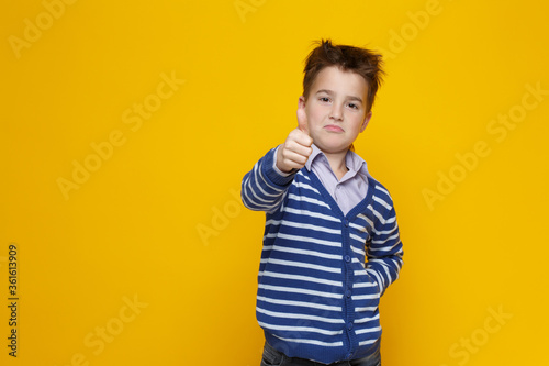 A little funny boy in a striped sweater shows the gesture of a raised thumb upwards on an outstretched arm isolated on a bright yellow background.