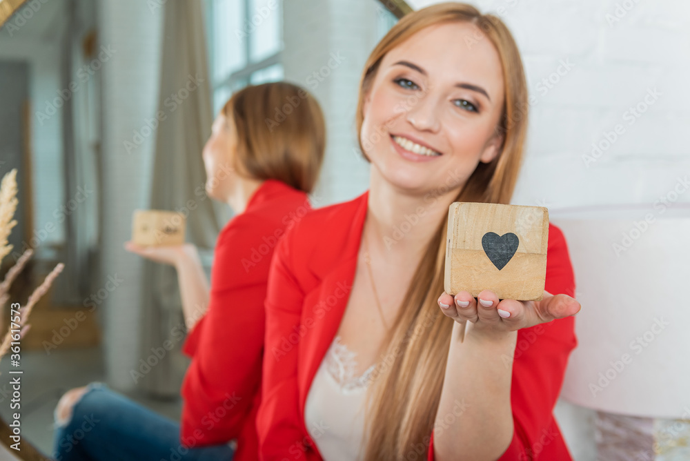 young pretty blonde girl in a red jacket sits near the mirror and holds a wooden cube with the image of a heart. The concept of motherhood young pretty blonde girl in a red jacket sits near the mirror and holds a wooden cube with the image of a heart. The concept of motherhood