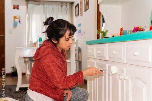 Close view of a young woman painting in white old furniture at home. Taken indoors under strong and bright white lights.