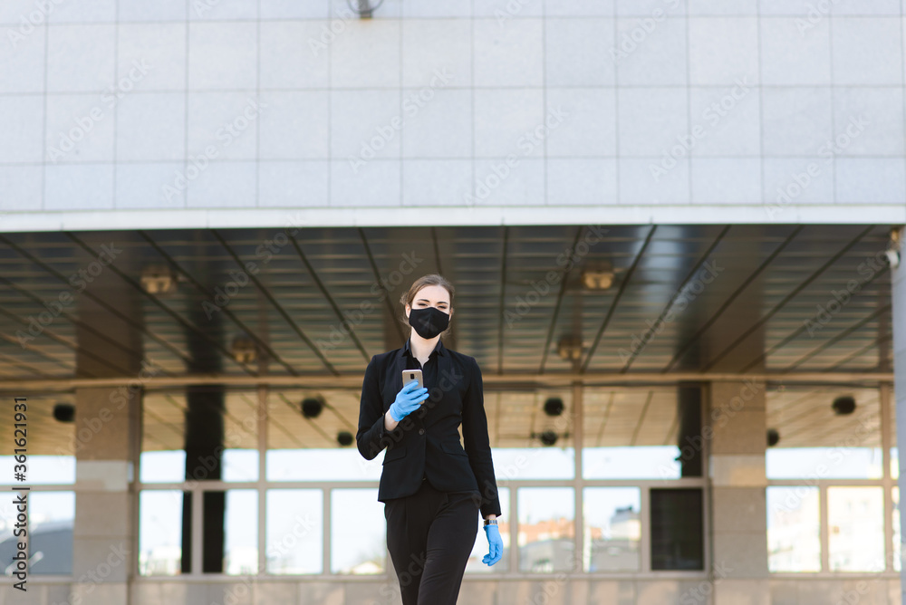 Beautiful businesswoman in a black suit in a black medical mask and gloves at the city in quarantine and isolation. Pandemic COVID-19. Selective focus Beautiful businesswoman in a black suit in a black medical mask and gloves at the city in quarantine and isolation. Pandemic COVID-19. Selective focus