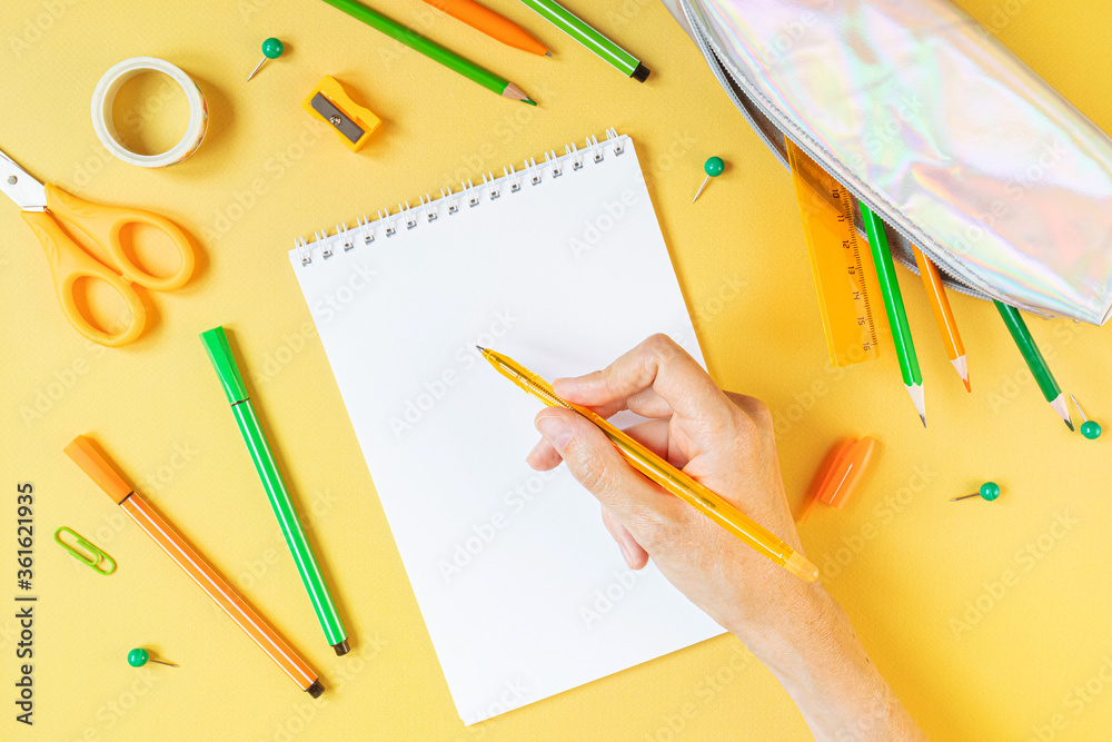 Concept back to school. School stationery flat lay: notebook, watercolor, pens, pencils, sharpener, scissors, ruler, paper clips on yellow background. Hand holds a pen. Orange and green tones. Concept back to school. School stationery flat lay: notebook, watercolor, pens, pencils, sharpener, scissors, ruler, paper clips on yellow background. Hand holds a pen. Orange and green tones.