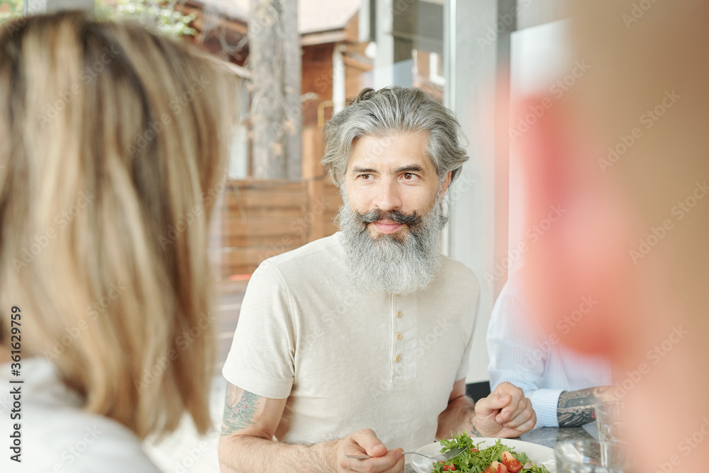 Handsome senior man with mustache and beard sitting at table and eating salad while talking to guests at dinner Handsome senior man with mustache and beard sitting at table and eating salad while talking to guests at dinner