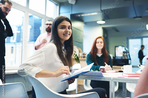 Young female manager writes important point of reference in time to the project meeting with colleagues. Attractive girl trainee keeps records for mastering new skills from the experts during briefing