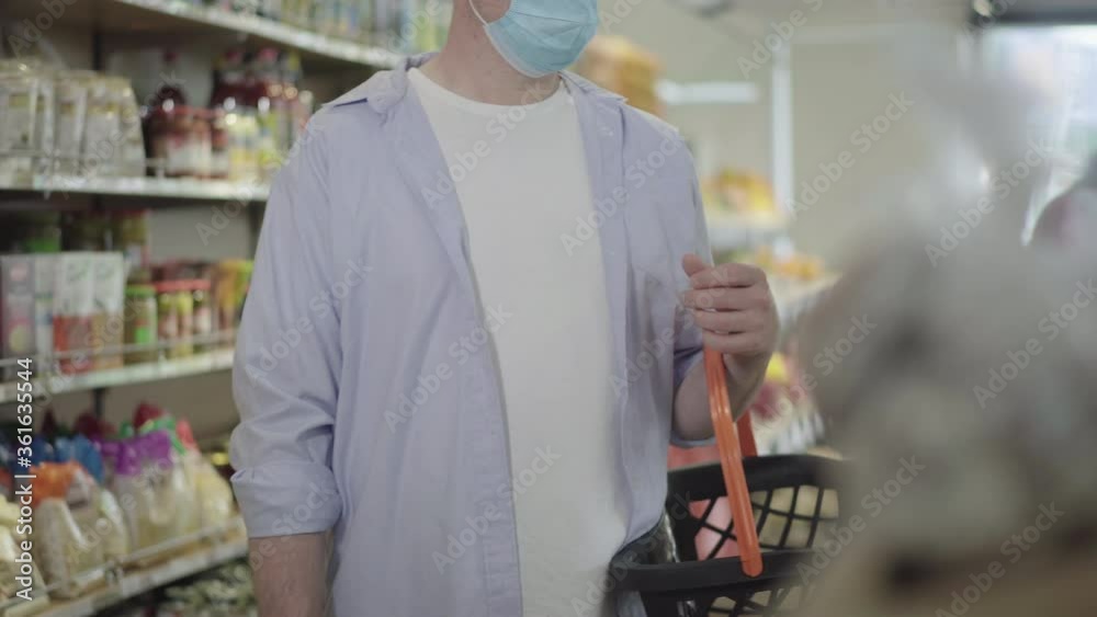 Unrecognizable man in face mask standing in grocery with shopping basket. Adult Caucasian male buyer purchasing good on Covid-19 pandemic in food shop. Coronavirus lifestyle.