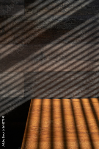 Light and shadow from the window. Thin lines of light on the wall and table. Shadow from the blinds. Brown wall and table in an empty room. 