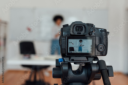 Young afro american woman standing near whiteboarad and recording English lesson on camera at home. Main focus on camera