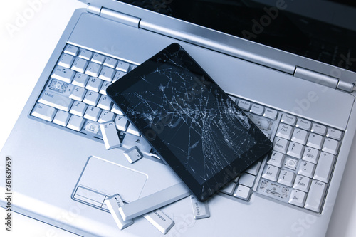 A silver laptop with a broken keyboard, tablet with a cracked display and black phone. A close-up picture of part of broken laptop and cracked screen on a tablet.