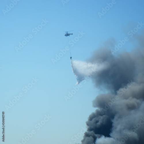 Aerial firefighting with helicopter. Black smoke from the fire over the city in the blue sky.