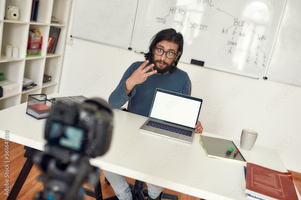 Fototapeta premium Young male teacher sitting at his workplace and explaining new theme while recording video blog on professional digital equipment. Coding class online