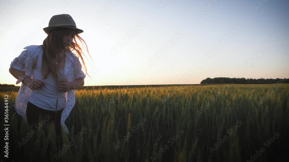 Pretty child in the hat is running across the wheat field. Happy young girl running in the field at sunset. Freedom concept.