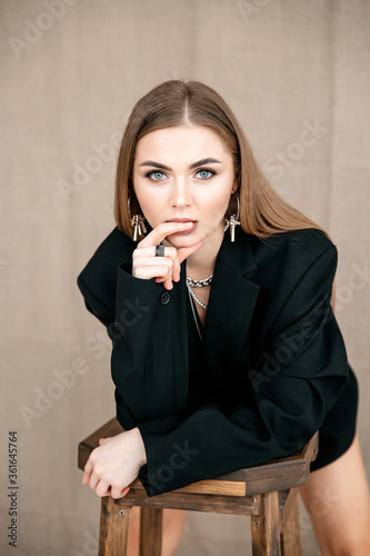 A beautiful girl of European appearance with brown hair, neutral makeup, in a black jacket looks into the frame with one hand near her face, a finger on her lips. Beige background.Vertical orientation