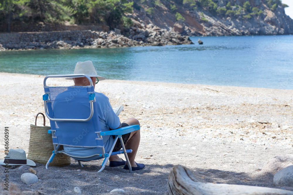 Fototapeta premium An older man in a hat sitting on a beach chair enjoying summer vacations on a Mediterranean sea beach. The man is on his back and has views of the sea