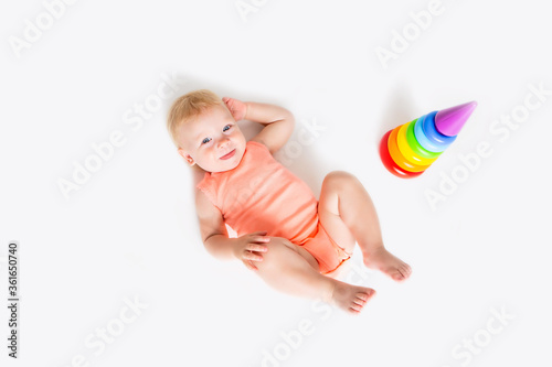 Toddler girl in orange bodysuit lies next to a multi-colored pyramid on a white background, view from above, space for text