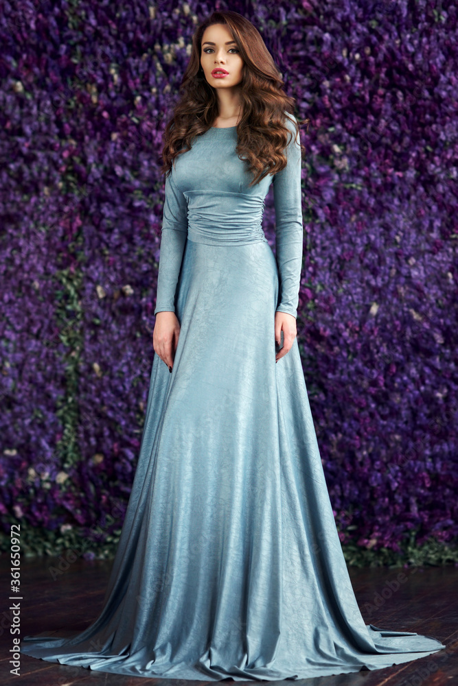 Young beautiful elegant woman in blue long evening dress posing against wall of purple flowers. Spring fashion style portrait of gorgerous girl Young beautiful elegant woman in blue long evening dress posing against wall of purple flowers. Spring fashion style portrait of gorgerous girl