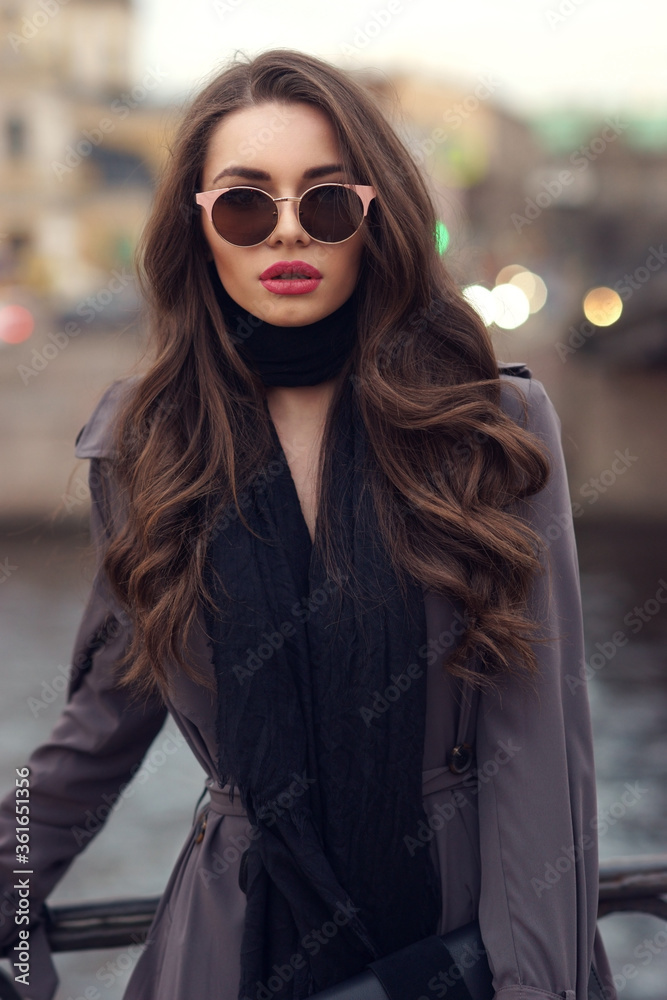 Fashionable glamorous girl with long curly hair wearing black dress, scarf, gray classy coat and sunglasses posing at city streets and looking at you or in camera. Fashion vogue style outdoor portrait Fashionable glamorous girl with long curly hair wearing black dress, scarf, gray classy coat and sunglasses posing at city streets and looking at you or in camera. Fashion vogue style outdoor portrait