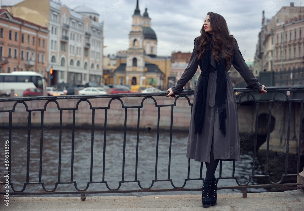 Young beautiful stylish girl with curly hair wearing black dress, scarf and gray coat standing near cast iron fence at city streets on a spring or atumn day
