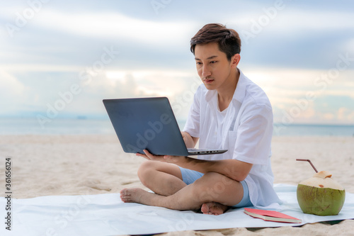 Young asian man working outdoor during his vacation time by laptop while sitting on the beautiful beach. Summer, holidays, vacation and happy people in Thailand concept..