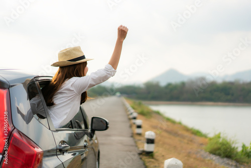 Woman relaxing and enjoying road trip. Young Asian happiness female wearing hat inside compact black car with raise her hand out to the car window with mountain and lake view.