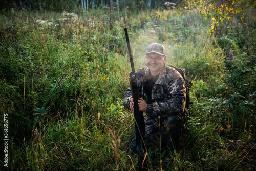 The smiling hunter or military person with gun or rifle in summer forest during the bird hunt.