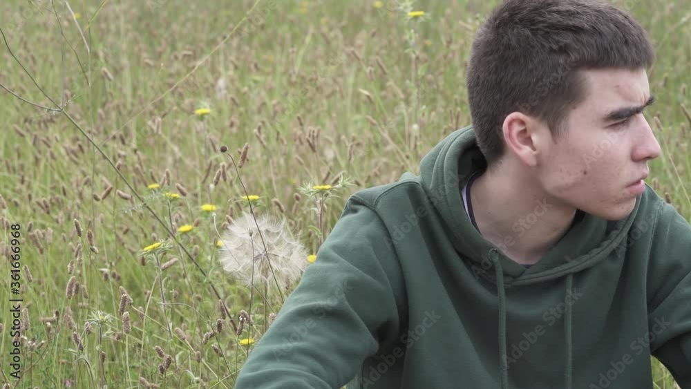 young guy sitting in a field and blowing out a dandelion young guy sitting in a field and blowing out a dandelion