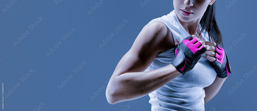 Conceptual close up portrait of fitness athletic young female model in  sports clothing. Confident female bodybuilder with power hand in gloves  over toned blue background Photos