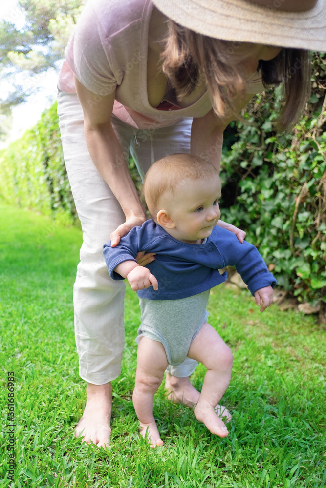 Lovely baby girl in blue shirt doing first steps with help of mom. Young unrecognizable mother holding daughter on grass. Closeup view. First barefoot steps, garden and sunny days concept Lovely baby girl in blue shirt doing first steps with help of mom. Young unrecognizable mother holding daughter on grass. Closeup view. First barefoot steps, garden and sunny days concept