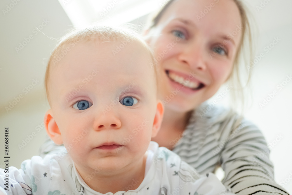 Serious adorable blue eyed baby looking at camera, blurred mom smiling in background. Closeup shot, front view. Child care or childhood concept Serious adorable blue eyed baby looking at camera, blurred mom smiling in background. Closeup shot, front view. Child care or childhood concept