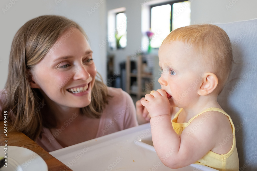 Joyful young mom watching baby daughter biting piece of food. Closeup shot. Child care or nutrition concept Joyful young mom watching baby daughter biting piece of food. Closeup shot. Child care or nutrition concept