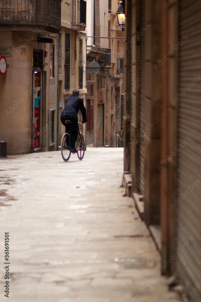A man is cycling in a narrow street among houses and shops on a cobblestone ground. Image was taken in old town in Barcelona, Spain A boutique as well as shutters of closed shops are in the background
