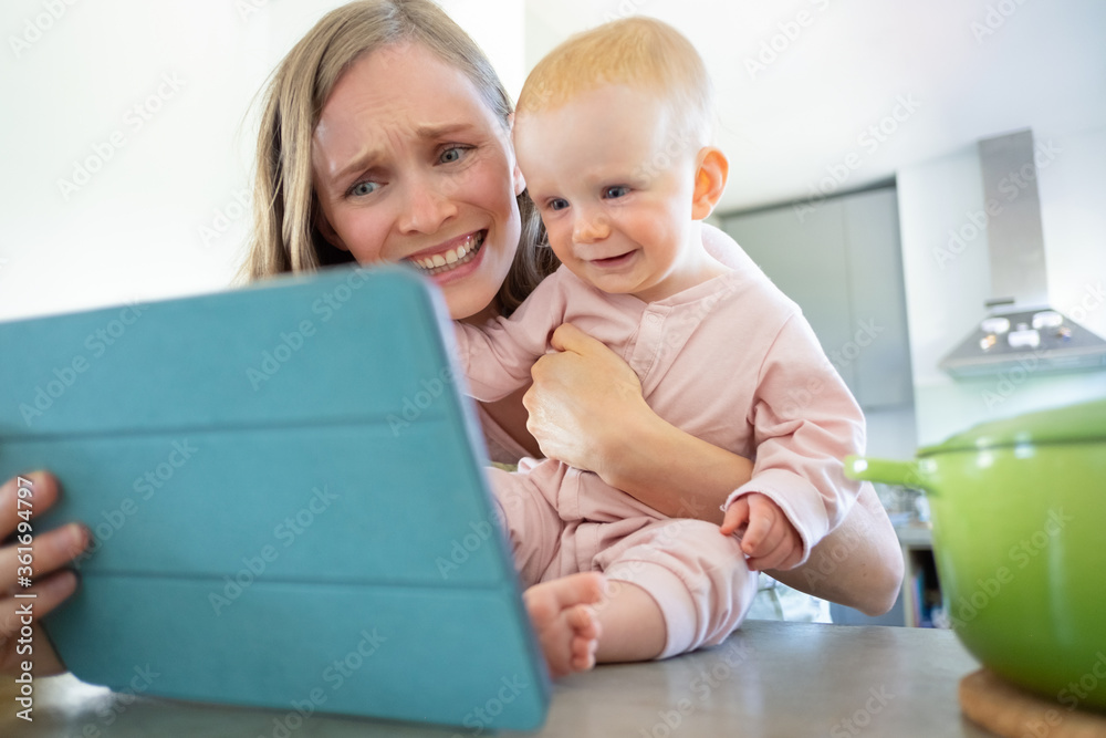 Cheerful mom and baby daughter laughing at tablet screen, watching online seminar or having video call. Child care or cooking at home concept Cheerful mom and baby daughter laughing at tablet screen, watching online seminar or having video call. Child care or cooking at home concept