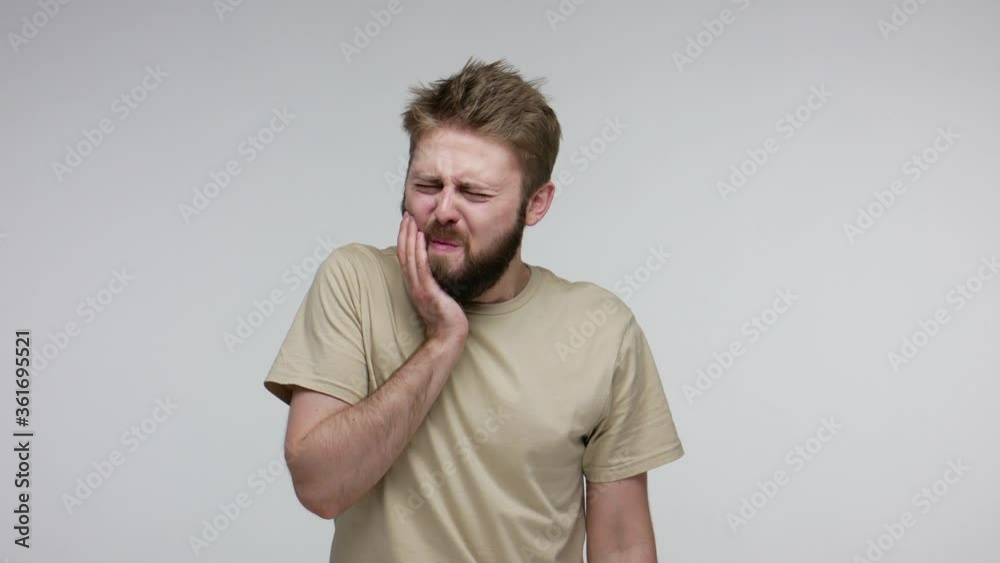 Dental problems. Unhealthy bearded man touching sore cheek frowning from pain, suffering toothache, cavities or gingivitis symptoms, gums disease. indoor studio shot isolated on gray background