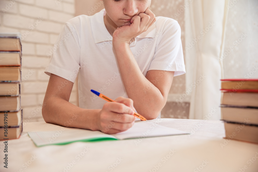 student sits at the table at the bookside with a pen in his hands. A tired teen does homework at home. Learning the concept. student sits at the table at the bookside with a pen in his hands. A tired teen does homework at home. Learning the concept.