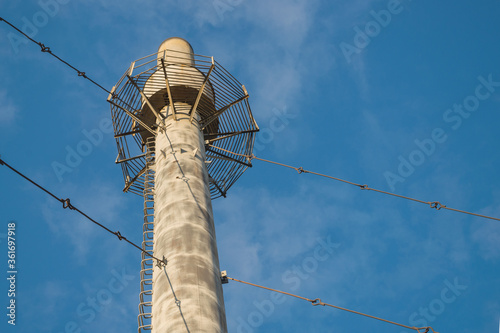 View from below of asbestos-cement boiler house pipe installed vertically, supported by system of metal tripwires, with observation platform at top and ladder of brackets rising up against cloudy sky photo