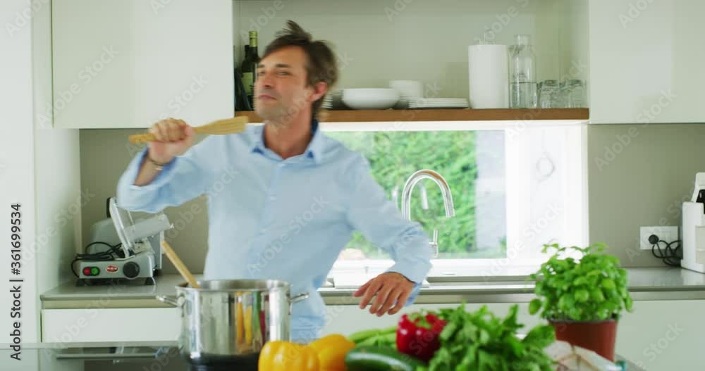 Authentic shot of a carefree happy smiling man is listening to the music and having fun to dance and sing with a wooden spoon during cooking a dinner in the kitchen. Authentic shot of a carefree happy smiling man is listening to the music and having fun to dance and sing with a wooden spoon during cooking a dinner in the kitchen.