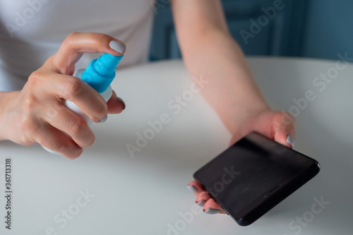 Woman spraying antiseptic  cleaning black smartphone on white table - close up view. Disinfection  protection  prevention  COVID 19  coronavirus  safety  sanitation  technology concept