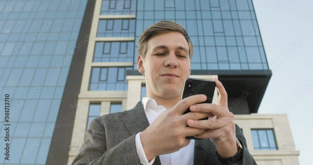 Young businessman is typing on smartphone near office building in downtown. Business, overwork, overtime working concept. Online job, study, modern life. Skyscraper on the background. Young businessman is typing on smartphone near office building in downtown. Business, overwork, overtime working concept. Online job, study, modern life. Skyscraper on the background.