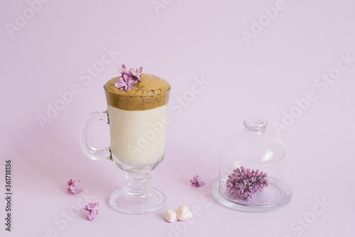 trendy drink 2020 dalgon Coffee in a transparent Cup with a paper tube, a plate with lilac flowers on a lilac background with a copy space photo