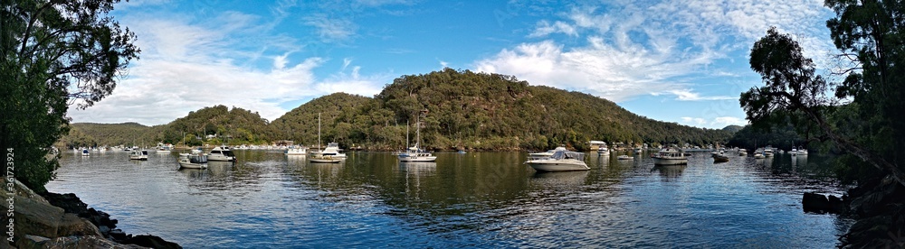 Fototapeta premium Beautiful morning panoramic view of Creek with boats and reflections of blue sky, light clouds, mountains and trees. Berowra Waters, Berowra Valley National Park, New South Wales, Australia