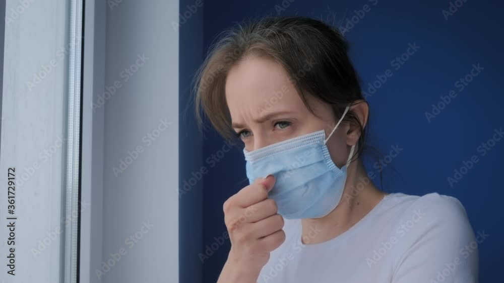 Slow motion: woman wearing face mask, feeling sick, suffering from coughing, looking out of window in room with blue wall. Self isolation, prevention, quarantine, COVID 19, coronavirus, safety concept Slow motion: woman wearing face mask, feeling sick, suffering from coughing, looking out of window in room with blue wall. Self isolation, prevention, quarantine, COVID 19, coronavirus, safety concept