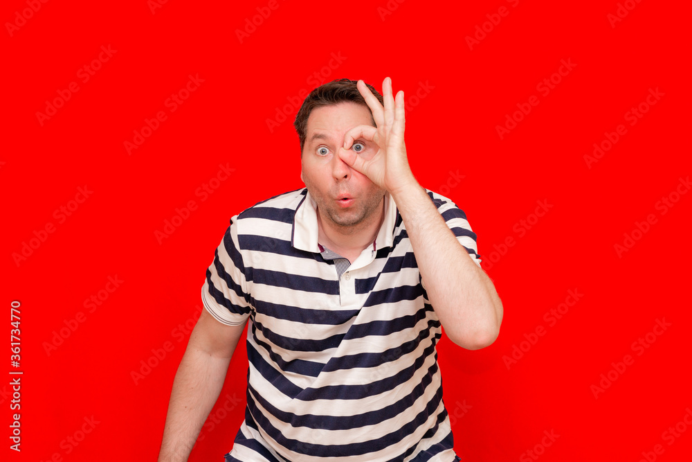 Joyful caucasian man wearing striped t-shirt standing over red surprised face, eye looking through fingers Joyful caucasian man wearing striped t-shirt standing over red surprised face, eye looking through fingers