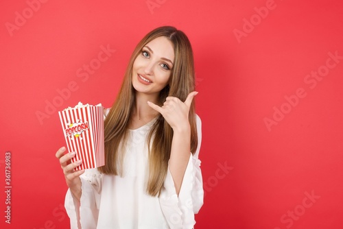 Young caucasian woman holding bucket with popcorn standing over isolated red background smiling doing phone gesture with hand and fingers like talking on the telephone. Communicating concepts.