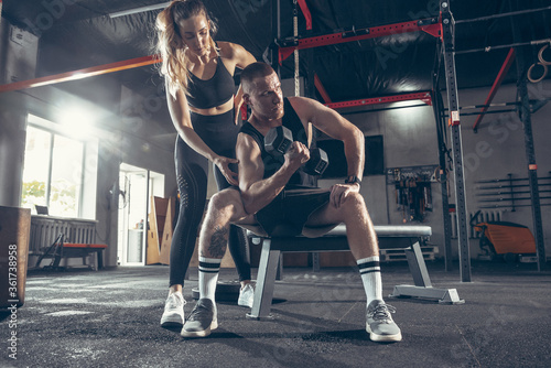Fototapeta Naklejka Na Ścianę i Meble -  Beautiful young sporty couple workout in gym together. Caucasian man training with female trainer. Concept of sport, activity, healthy lifestyle, strength and power. Working out with weights.
