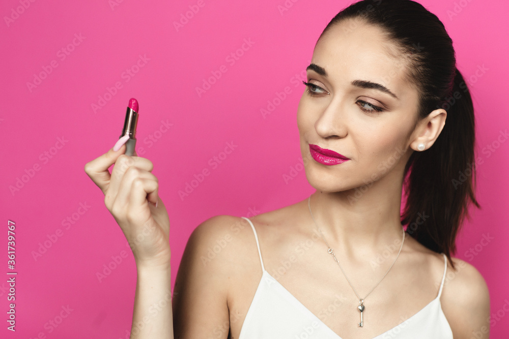 Caucasian woman with a ponytail on a pink background. Young woman with pink lips looking at lipstick. Model posing in the studio, advertises cosmetics Caucasian woman with a ponytail on a pink background. Young woman with pink lips looking at lipstick. Model posing in the studio, advertises cosmetics